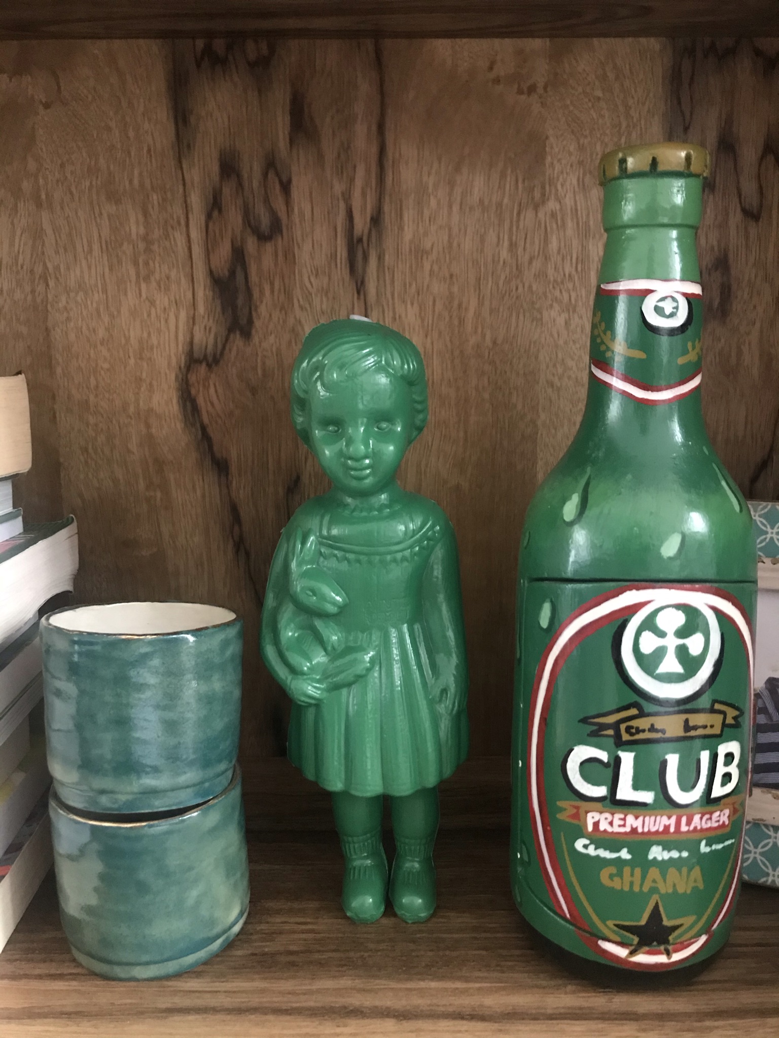 A green plastic Clonette Doll stands on our bookshelf next to a wooden mimic of a Ghanaian fantasy coffin in the shape of a Club beer bottle.