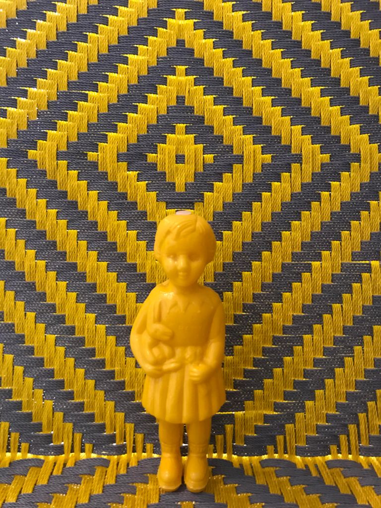 Yellow baby Clonette Doll stands against a backdrop of a yellow and grey chair woven by Joel of Calao Creations in Ghana